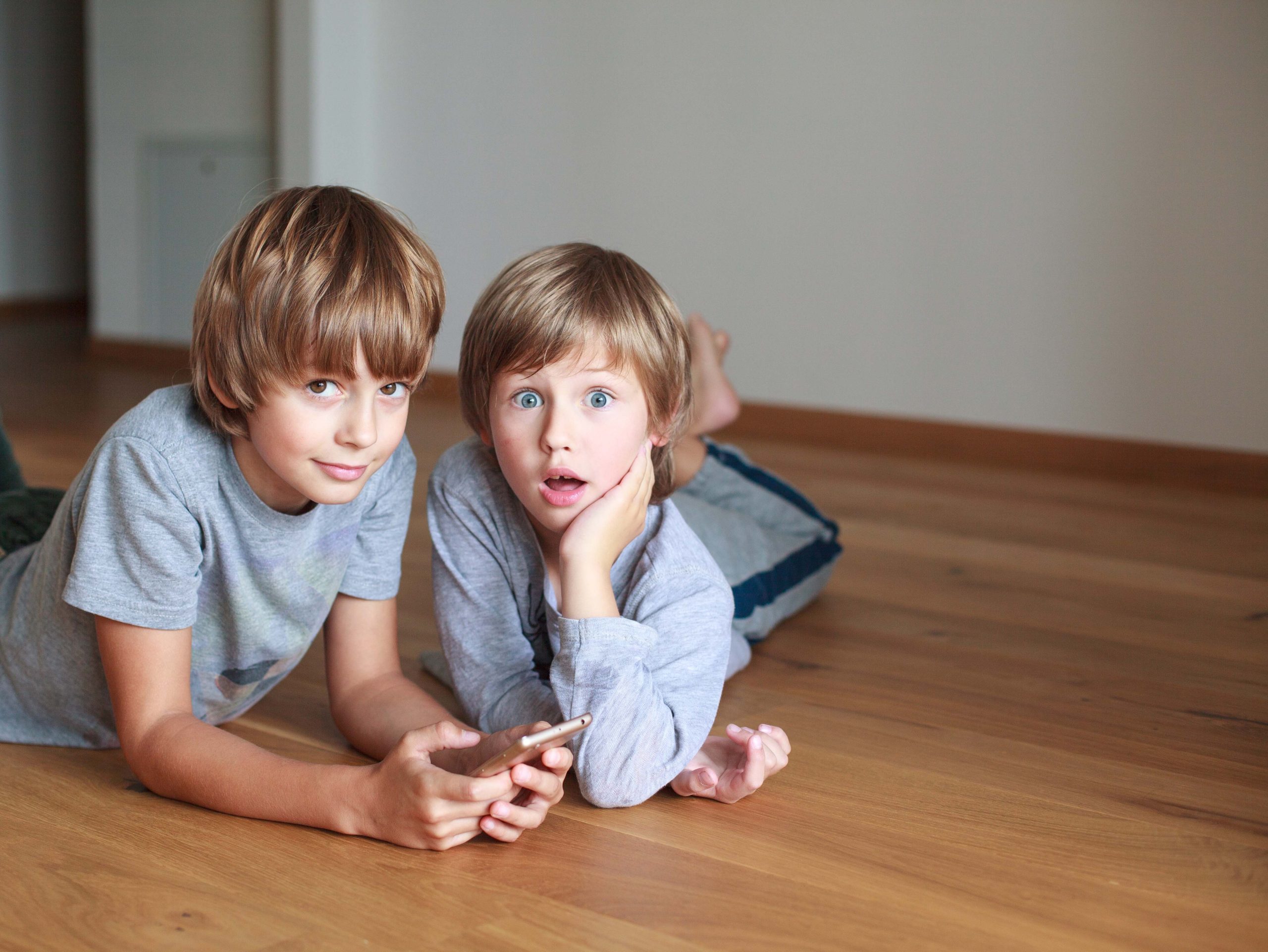 Two surprised kids lying on wooden floor and using mobile phone